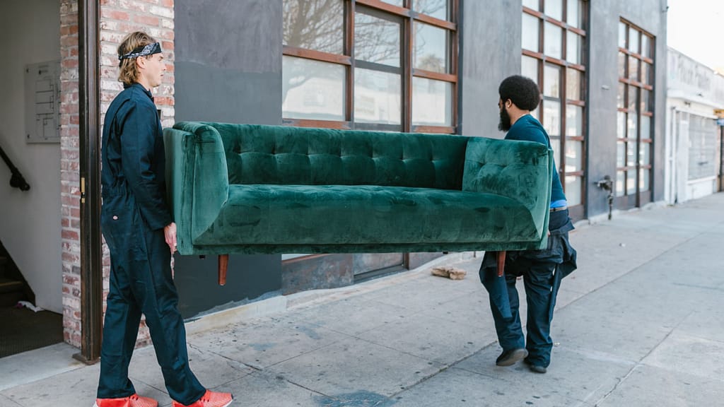 Two movers carrying a green velvet sofa along a sidewalk, part of long distance moving tips for furniture
