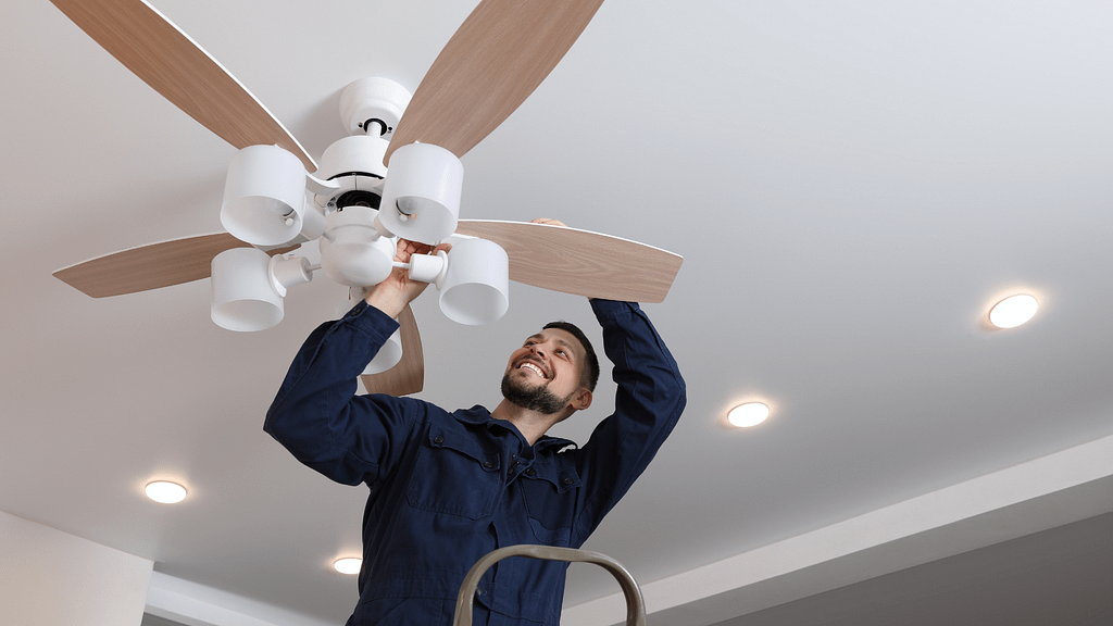 Man on a ladder installing a wood-blade ceiling fan on a white ceiling with recessed lights